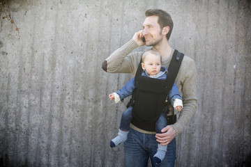 Young man using mobile phone while carrying baby boy in carrier against wall