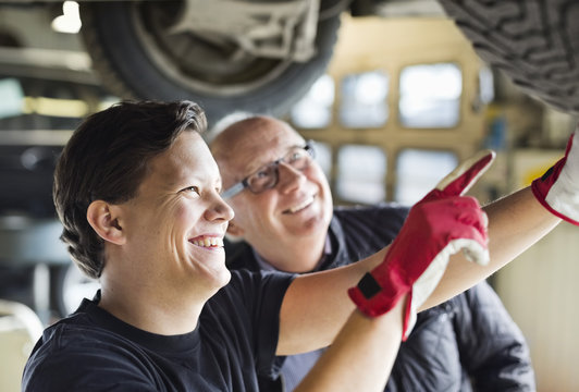 Happy young mechanic explaining tire problems to customer underneath car at auto repair shop