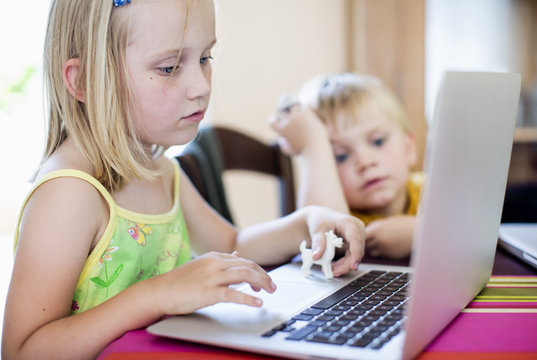 Girl using laptop at table with brother sitting in background