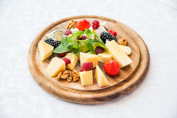 разные сорта сыров с ягодами ежевики, малины и земляники, украшенные мятной веточкой на круглой доске и белой скатерти 