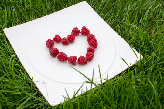 Fresh red raspberries in a heart shape on a white plate, green grass background