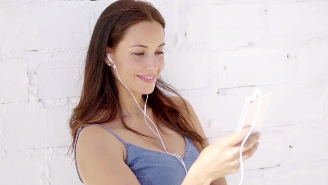 Attractive young long haired brunette woman standing in front of a white brick wall listening to music on earphones
