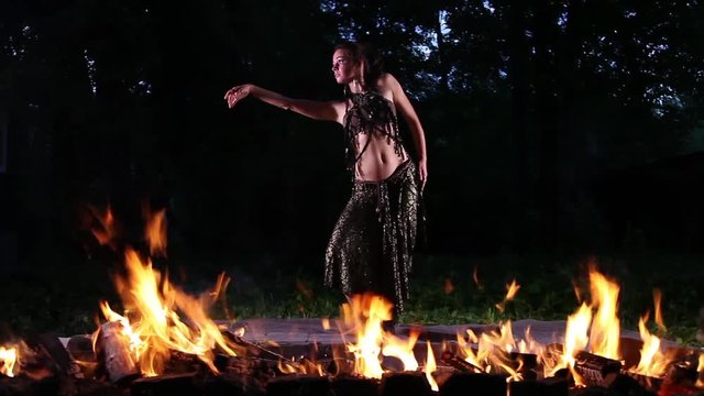 Pretty young woman dancing in the night with a fire flame in the foreground
