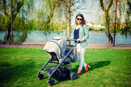 Trendy young mother smiling and walking with baby in pram in park