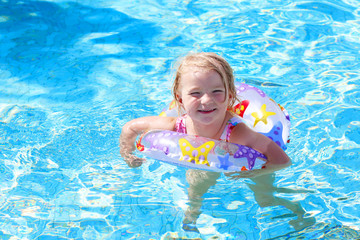 Adorable happy little child, curly toddler girl in swimming suit having fun relaxing and floating on an inflatable toy ring in a pool on sunny day during summer vacation in resort
