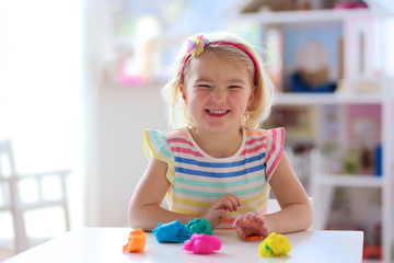 Little preschooler girl playing with plasticine. Happy creative toddler girl playing with dough, colorful modeling compound, sitting at white table in sunny room at home or kindergarten