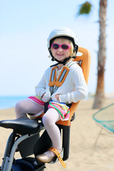 Naklejka premium Happy little girl enjoying ride on the bike. Toddler sitting in child bicycle seat. Family having fun outdoors. Beach promenade and sea at the background.