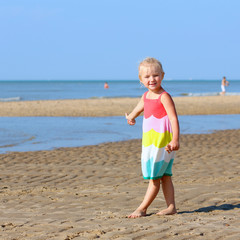 Cute active child wearing colorful dress playing on wide sandy beach. Happy little girl enjoying summer holidays on a sunny day. Family with young kids on vacation at the North Sea coast.