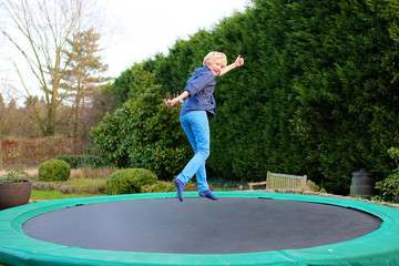Happy kid plays outdoors in garden jumping high in the sky on trampoline. Active teenager boy having fun outdoors at early spring day. Healthy lifestyle concept.