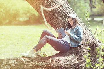 Happy young female student is reading in a park. She is sitting on a tree and enjoying her book.