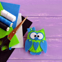 Cute felt owl ornament. Owl is sewn from green and blue felt and decorated with small buttons. Hand fabric embellishment idea. Felt kit, thread, needle, scissors on lilac wooden background
