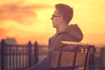 Fototapeta na wymiar Photo of Lonely Man Sitting on a Bench during Sunset. Portrait of Young Man Sitting on Wooden Bench. Sunset Male Outdoor Portrait