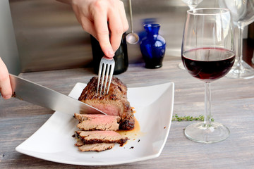 Just cooked roast beef is sliced by young chef on the white plate. Knife, fork, red wine, wine glass, wooden table
