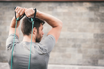 urban fitness man looking aside while working out  triceps using expander. Sporty strong male exercising outdoors with elastic straps.