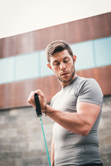 urban fitness man looking at his biceps while working out arms using expander. Sporty strong male exercising outdoors with elastic straps.