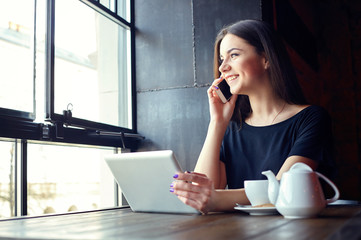 Young attractive girl talking on mobile phone and smilling while sitting alone in coffee shop during free time and working on tablet computer. Happy female having rest in cafe. Lifestyle, coffee