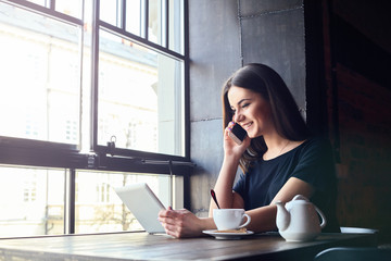 Young attractive girl with cute smile talking on mobile phone while sitting alone in coffee shop during free time and working on tablet computer. Happy female having rest in cafe. Lifestyle, coffee