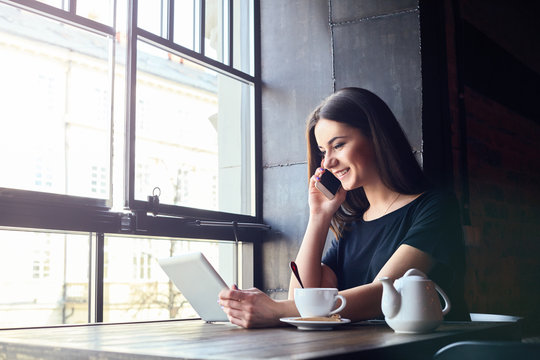 Young Attractive Girl With Cute Smile Talking On Mobile Phone While Sitting Alone In Coffee Shop During Free Time And Working On Tablet Computer. Happy Female Having Rest In Cafe. Lifestyle, Coffee