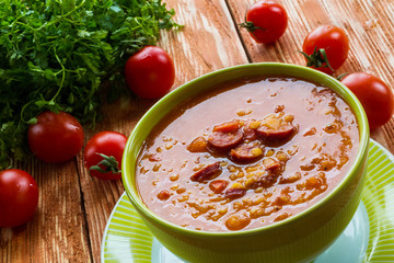 Soup: tomato soup with smoked sausage, tomatoes and lentil. Green bowl on wooden table. Small cherry tomatoes and green lettuce on table.