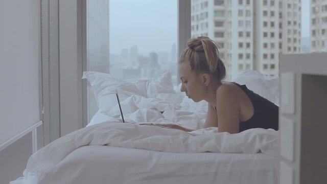 Elegant, smart, young casual looking woman using her tablet  computer  or laptop in bed in a luxury apartment.