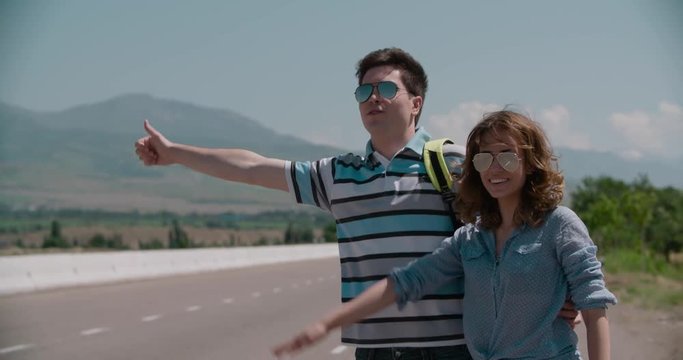young couple standing on an empty road, hitchhiking, mountainous terrain, slow motion