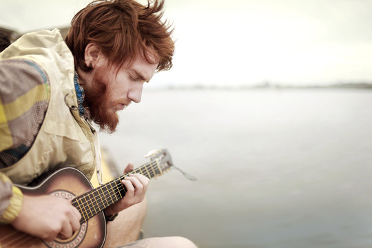 Man playing guitar while sitting by river against sky