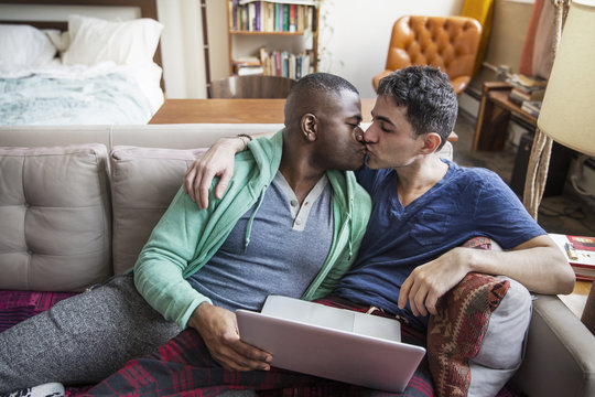 Gay couple kissing each other while holding laptop at home