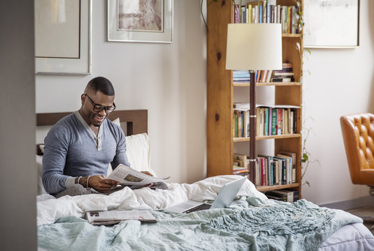 Smiling young man reading newspaper while sitting on bed