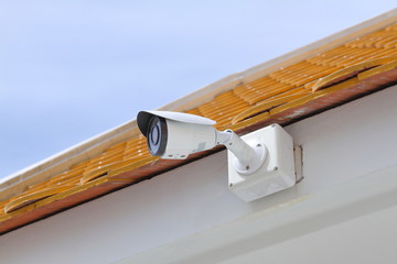 CCTV security camera attached to white wall under Thai Buddhism temple yellow, orange roof top scale