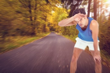 Tired athlete wiping his sweat with hand against country road along trees in the lush forest