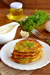 Vegetable fritters cooked with zucchini, garlic and dill. A stack of zucchini fritters on a plate. Bottle with vegetable oil, gravy with sour cream, garlic, green dill, fork on a wooden table. Macro