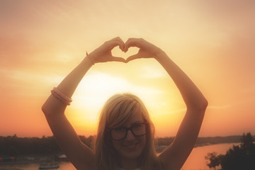 Girl holding a heart-shape with sunset / sunrise background.