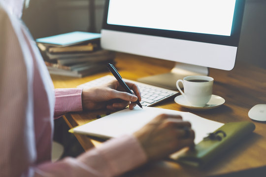 Young girl writing in a notebook in the office atmosphere on the keyboard and cup of coffee on the wooden table background, digital in female hands in the night, workspace