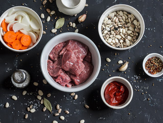 Raw ingredients - beef, beans, onions, carrots, spices, tomato sauce on a dark stone background