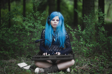 Young girl with blue hair playing synthesizer dressed in black sitting on grass in spring in park. Female composer and musician. Creative woman