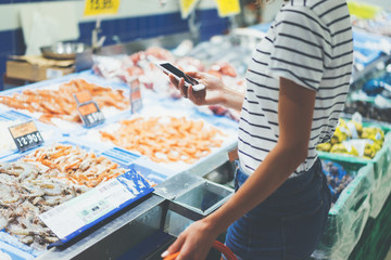 Young woman shopping healthy food in supermarket blur background. Close up view girl buy products using smartphone in store. Hipster at grocery using smartphone. Person comparing the price of produce.