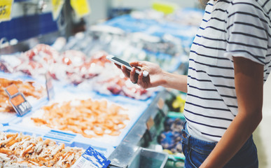 Young woman shopping healthy food in supermarket blur background. Close up view girl buy products using smartphone in store. Hipster at grocery using smartphone. Person comparing the price of produce.