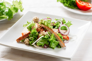 Plate with chicken salad and mushrooms closeup. Potion of salad with fried chiken and mushrooms on white background. Square white plate with chicken salad