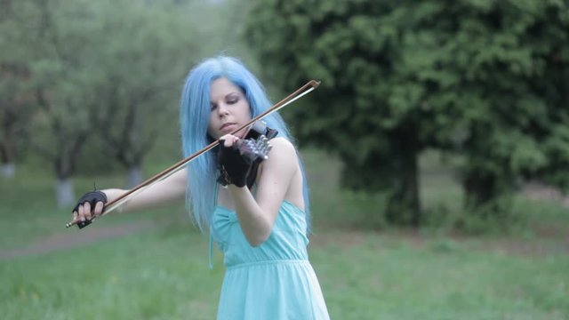 Young girl with blue hair playing violin in blue dress in spring in park. Female violinist, composer and musician. Creative woman