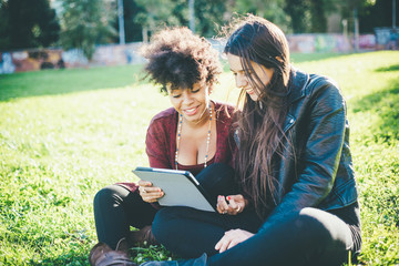 two multiethnic beautiful young woman black and caucasian using tablet sitting in a city park, lookind down the screen smiling - technology, social network, happiness concept