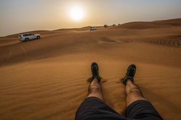 Watching sunset in Dubai sitting on dunes - point of view legs and safari jeeps