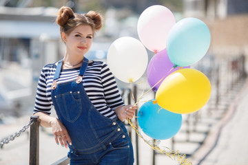 pregnant brunette woman with fashionable hairstyle, dressed in blue denim overalls and a striped shirt,posing with a bunch of colorful balloons on the pier on the background of boats and yachts