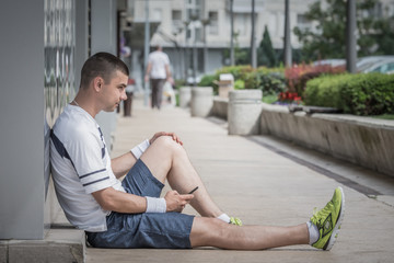 Obraz premium Sportsman resting by sitting against the wall and holding smart phone. Urban scene in the background. Urban and healthy lifestyle concepts. 