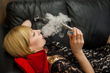 девушка на черном кожаном диване курит электронную сигарету . Girl on a black leather couch is smoking an electronic cigarette - obrazy, fototapety, plakaty