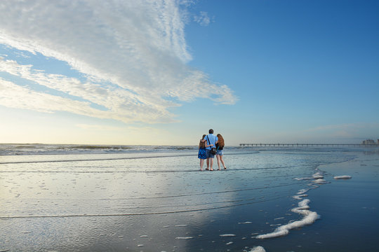 Family Enjoying Time Together On Beautiful Beach.Father With Arms Around His Daughters .Beautiful Cloudy Sky Reflected On The Beach, Pier In The Background. Copy Space. .Jacksonville, Florida, USA. 