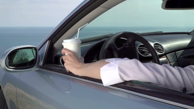 Relaxed young woman driver holding paper coffee cup in her car parked on the edge of cliff by sea businesswoman looking away beyond the horizon, close-up pan shot