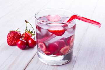 A glass of water with fruit, next to strawberries and cherries, tube on a wooden table
