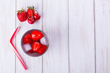 A glass of water with fruit, next to strawberries and cherries, tube on a wooden table