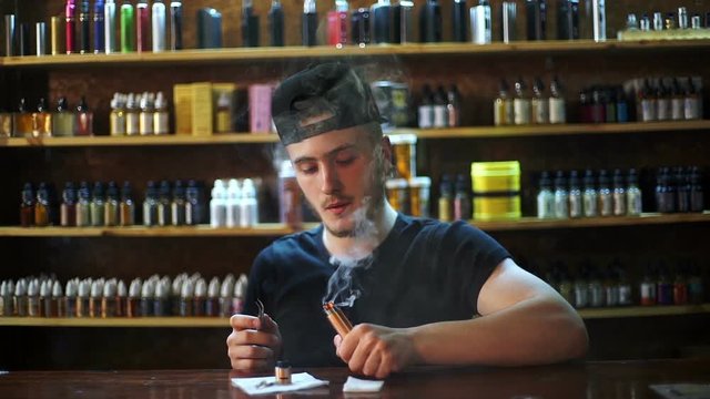 young seller in vape shop is installing cotton in atomizer show window in background