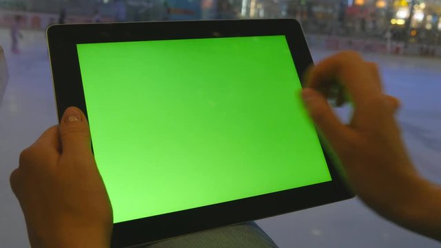 Female hands using tablet pc with green screen in shopping mall. Lot of people skate on indoor ice rink at the background
